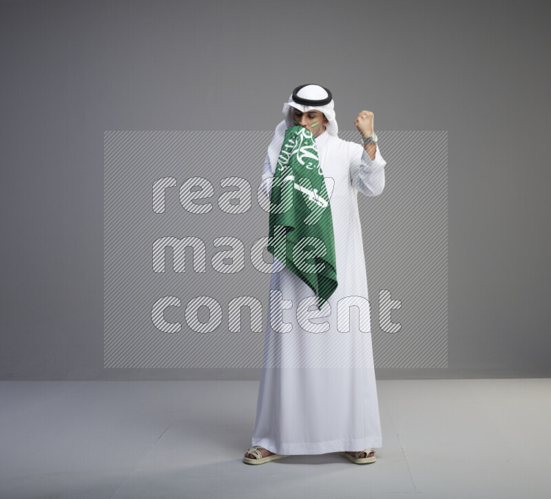 A Saudi man standing wearing thob and white shomag with face painting kissing big Saudi flag on gray background