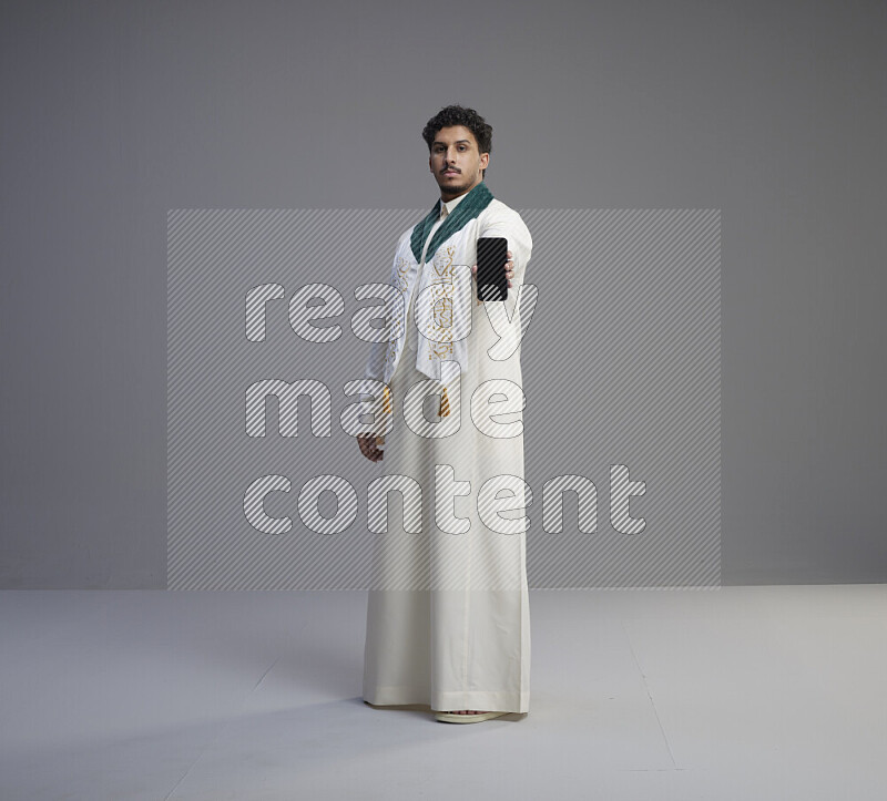 A Saudi man standing wearing thob and Saudi flag scarf showing phone to camera on gray background