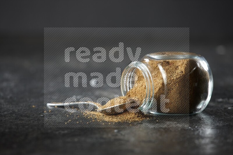 A flipped glass spice jar and a metal spoon full of cumin powder and powder spilled out on a textured black flooring