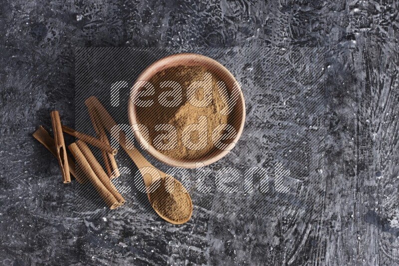 wooden bowl full of cinnamon powder and a wooden spoon full of it with cinnamon sticks on a textured black background