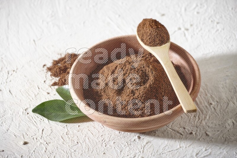 A wooden bowl and a wooden spoon full of cloves powder on a textured white flooring