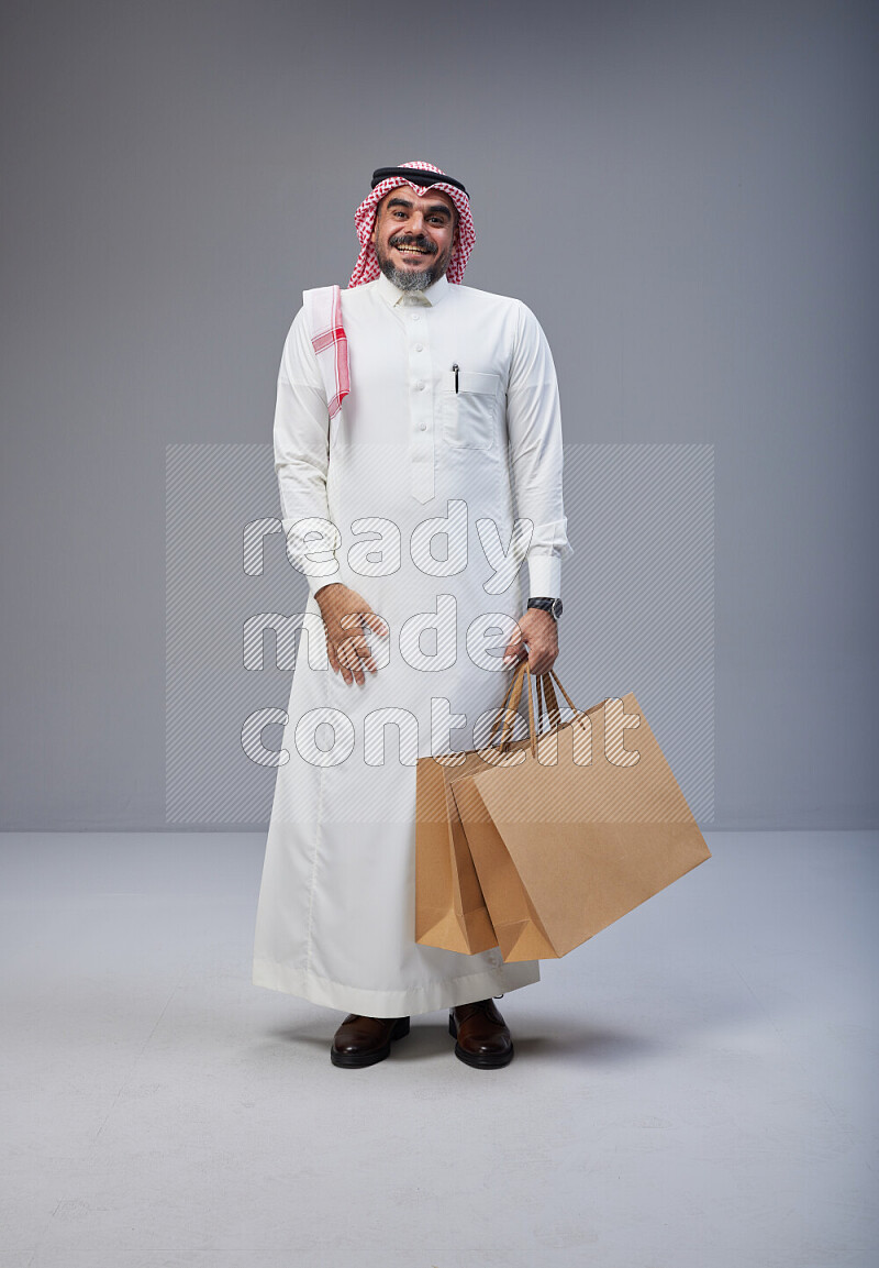 Saudi man Wearing Thob and red Shomag standing holding shopping bag on Gray background