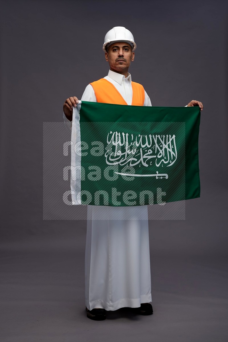 Saudi man wearing thob with engineer vest standing holding Saudi flag on gray background