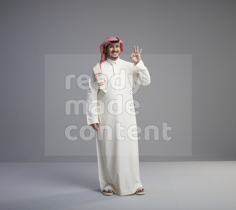 A Saudi man standing wearing thob and red shomag interacting with the camera on gray background