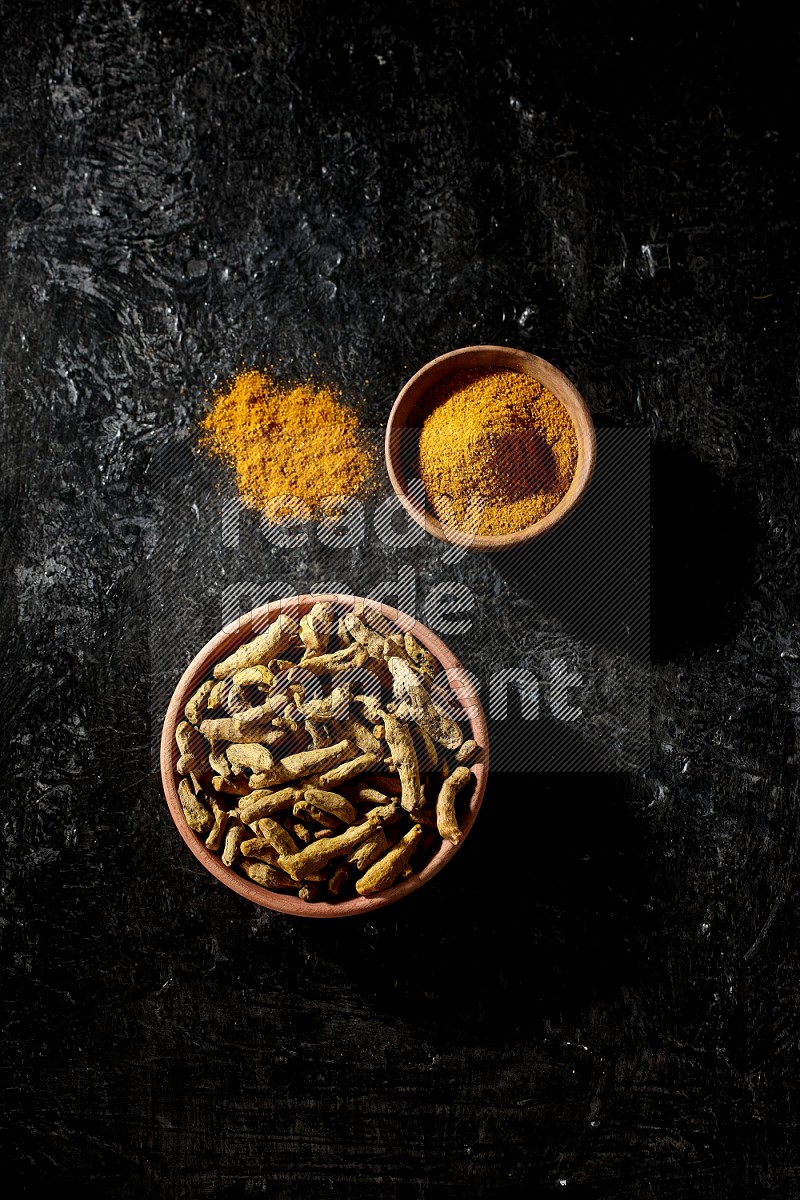 2 wooden bowls, one full of turmeric powder and the other full of dried turmeric whole fingers on a textured black flooring