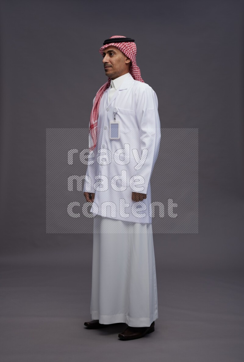 Saudi man wearing thob with lab coat and shomag with pocket employee badge standing interacting with the camera on gray background