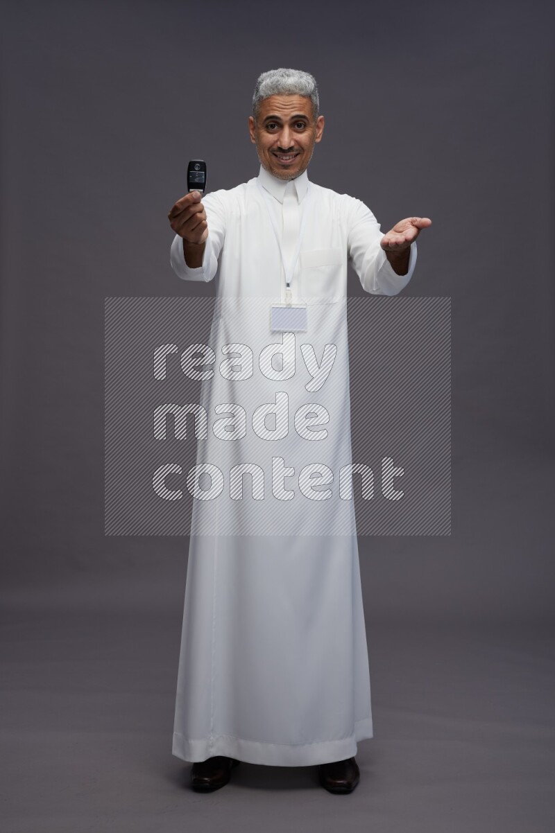 Saudi man wearing thob with neck strap employee badge standing holding key car on gray background
