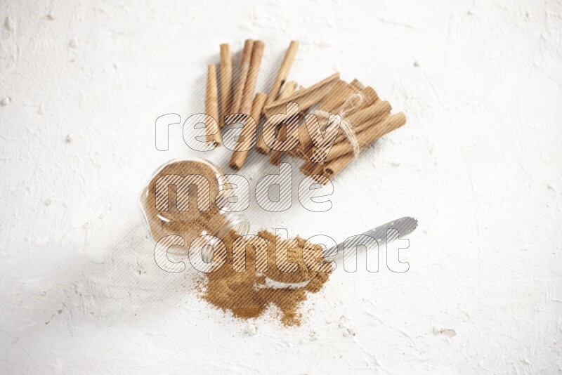 Flipped herbs glass jar full of cinnamon powder with a metal spoon full of powder and cinnamon sticks on a textured white background