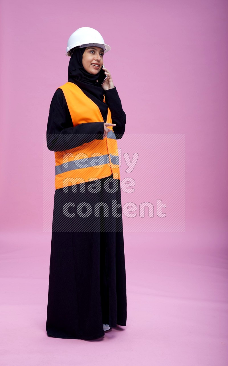 Saudi woman wearing Abaya with engineer vest and helmet standing talking on phone on pink background