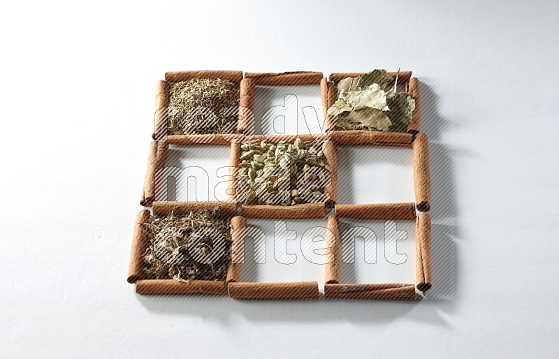 9 squares of cinnamon sticks full of cardamom in the middle surrounded by nutmeg, cinnamon, bay laurel leaves, cloves, cumin, dried ginger, dried basil and star anise on white flooring