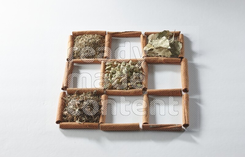 9 squares of cinnamon sticks full of cardamom in the middle surrounded by nutmeg, cinnamon, bay laurel leaves, cloves, cumin, dried ginger, dried basil and star anise on white flooring