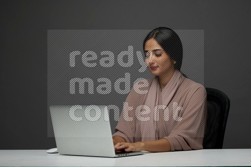 A Saudi woman Sitting on her desk on a Gray Background wearing Brown Abaya