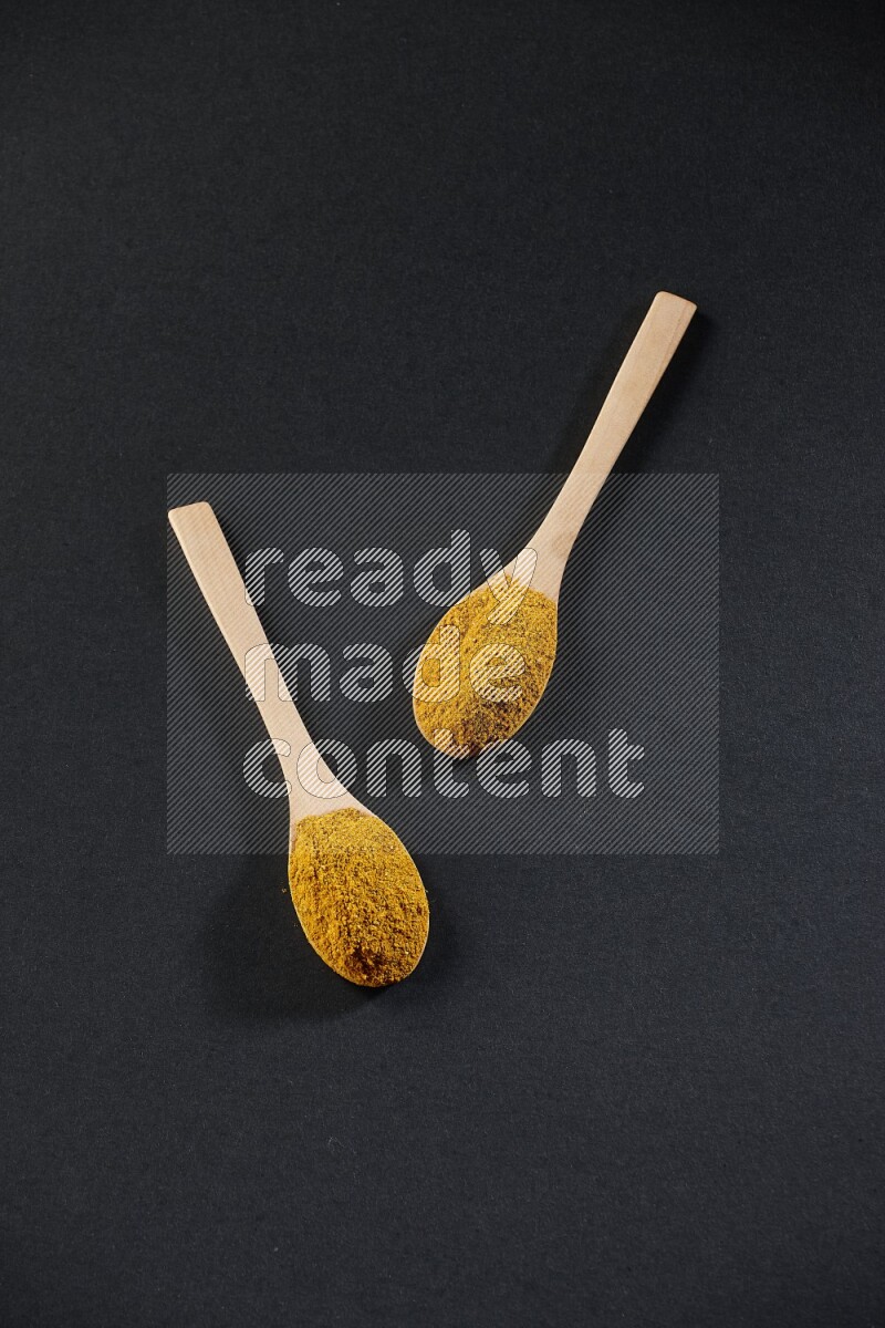 2 wooden spoons full of turmeric powder on black flooring