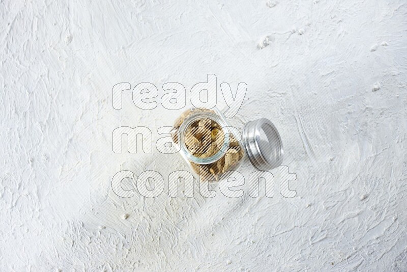 A glass spice jar full of dried turmeric whole fingers on a textured white flooring