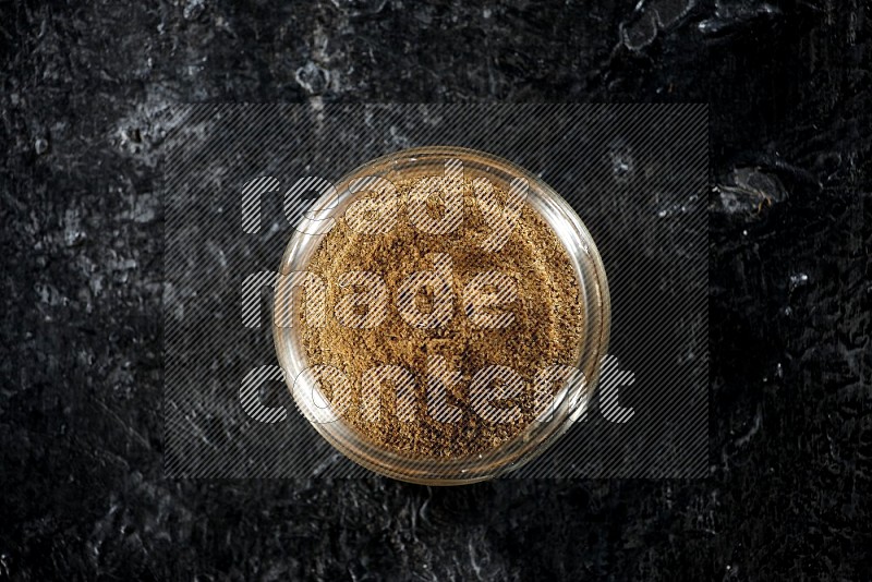 A glass jar full of cumin powder on a textured black flooring