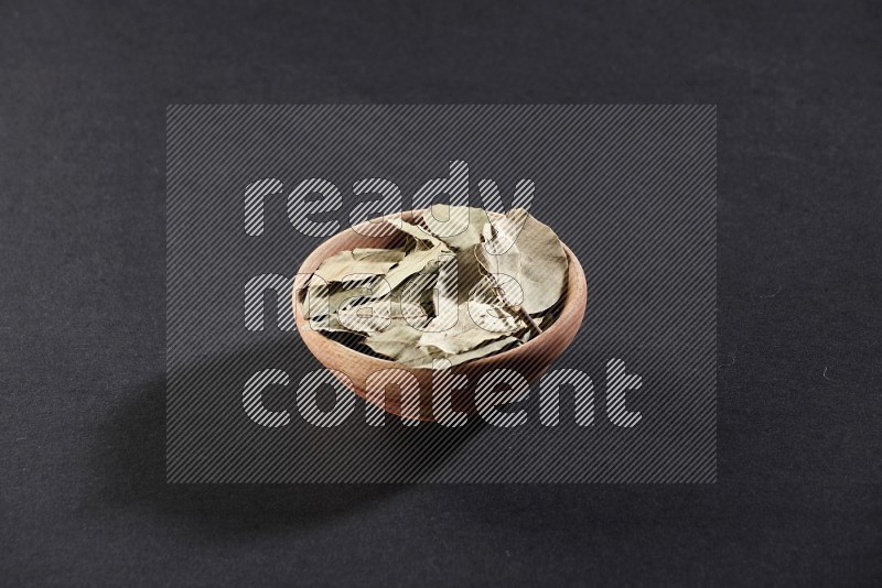 A wooden bowl filled with dried bay leaves on black flooring