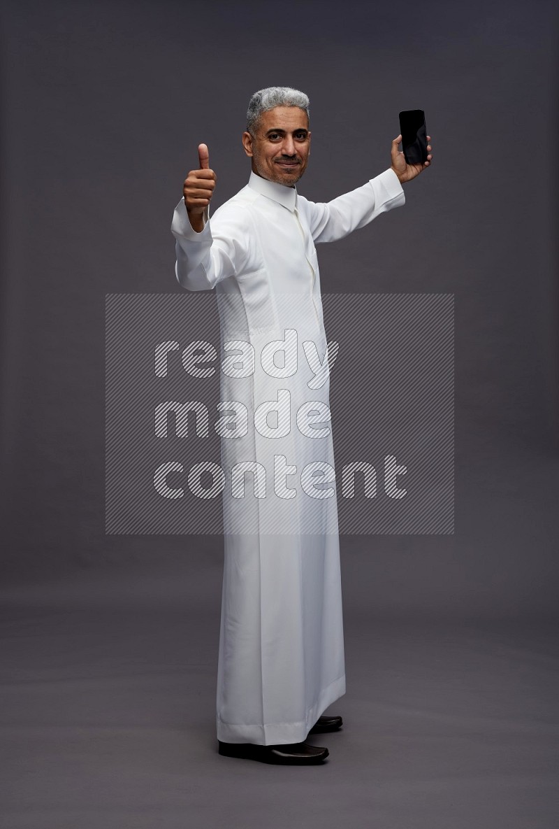 Saudi man wearing thob standing taking selfie on gray background
