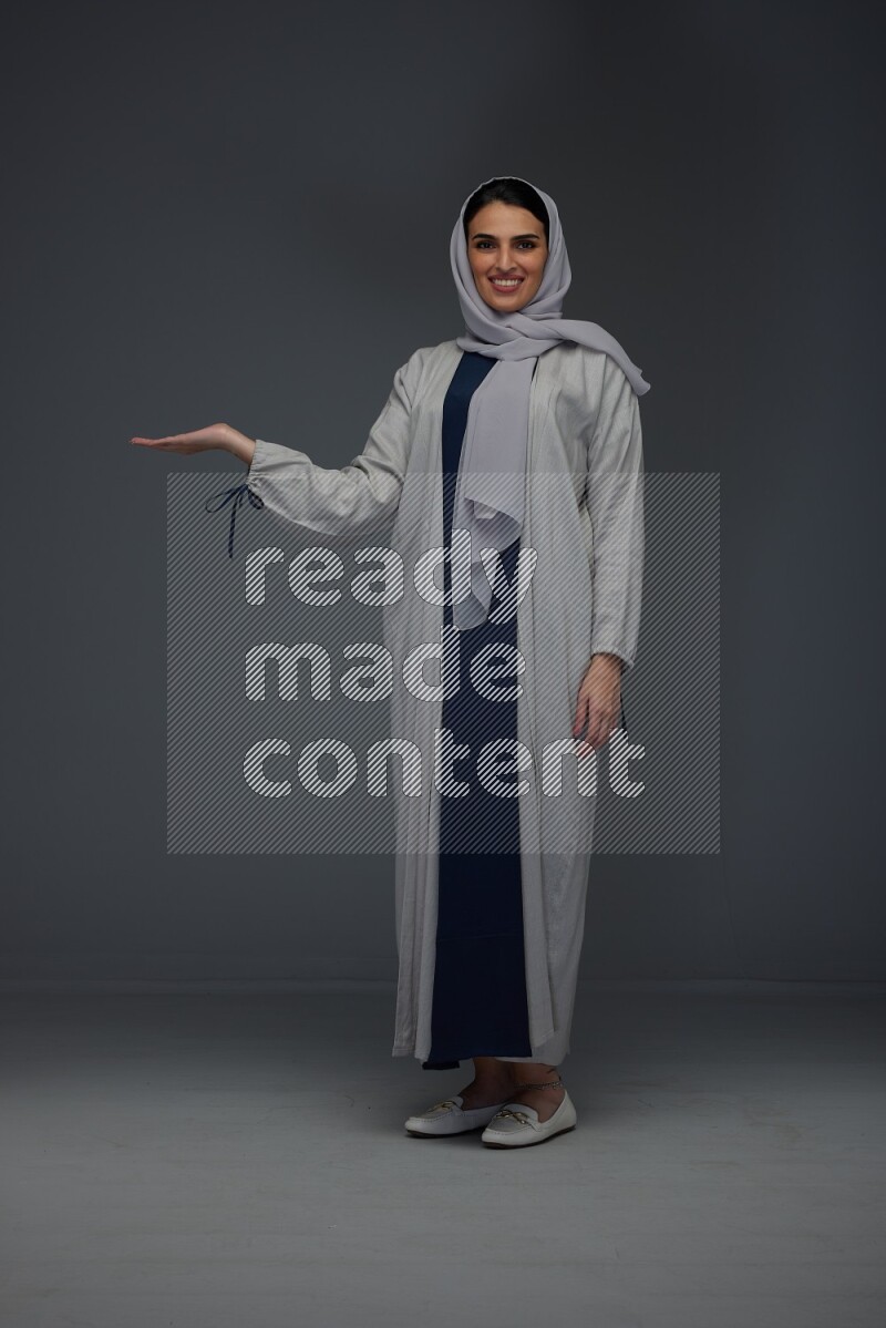 A Saudi woman wearing a light gray Abaya and head scarf standing and pointing in different directions eye level on a grey background