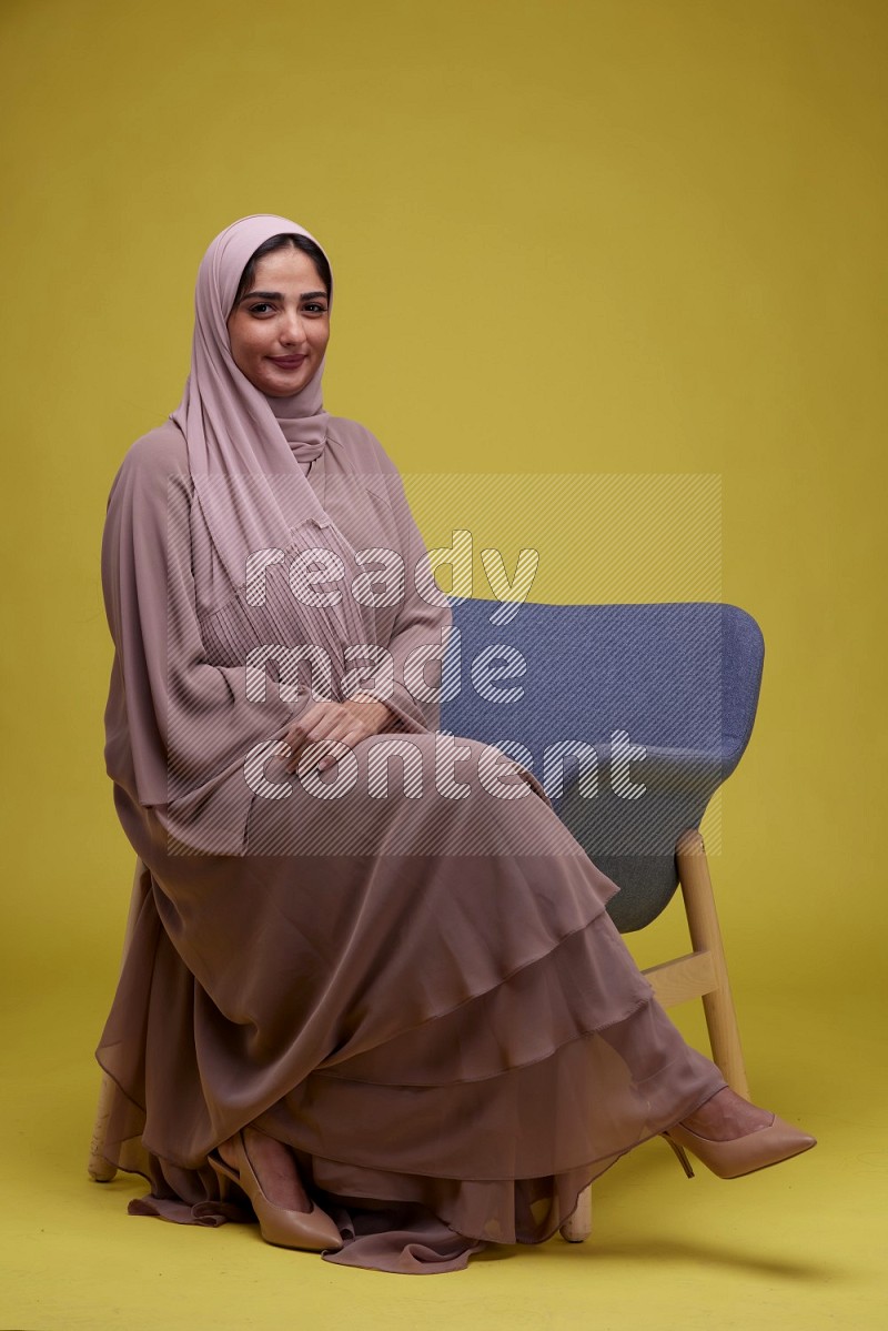 A Female sitting on a Blue Chair on a Yellow Background wearing Brown Abaya with Hijab