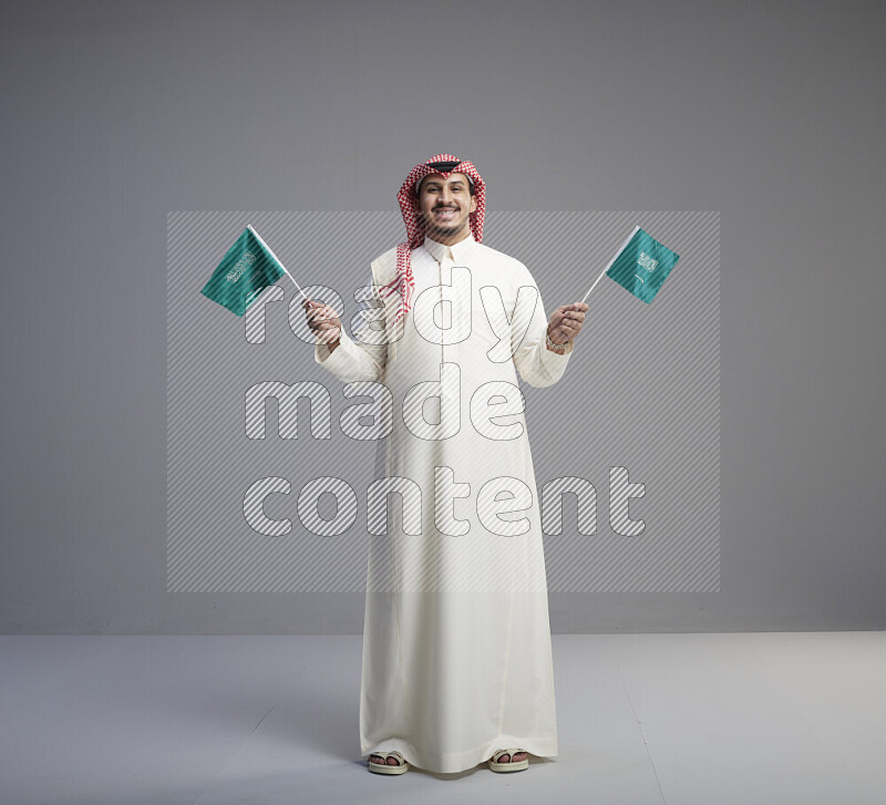 A Saudi man standing wearing thob and red shomag raising small Saudi flag on gray background