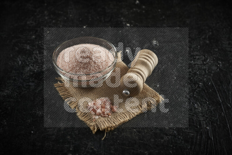 A glass bowl full of pink himalayan salt with a wooden grinder on a burlap fabric all on black background