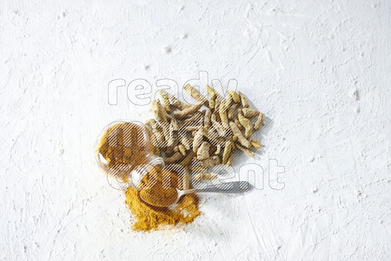 A flipped glass spice jar and metal spoon full of turmeric powder and powder spilled out of it with dried whole fingers on textured white flooring
