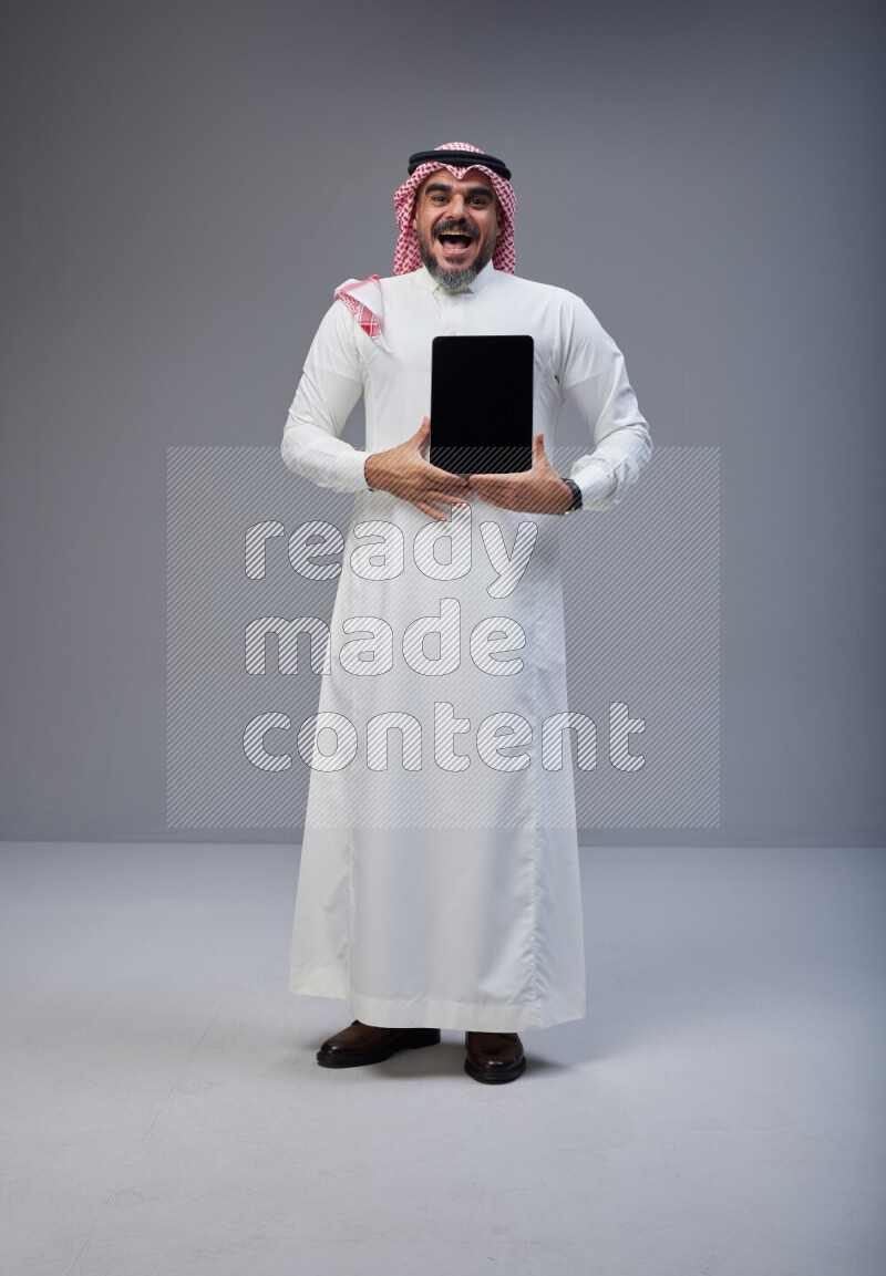 Saudi man Wearing Thob and red Shomag standing showing tablet to camera on Gray background