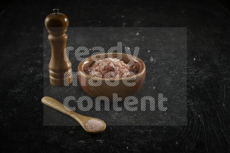 A wooden bowl and spoon filled with coarse pink himalayan salt and a wooden grinder beside them on black background