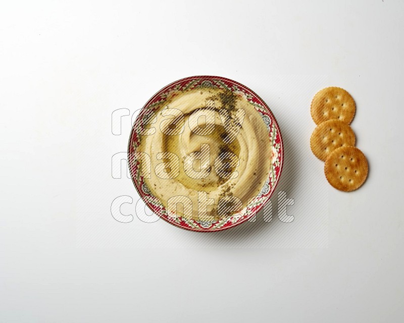 Hummus in a red plate with patterns garnished with zaatar & sumak on a white background