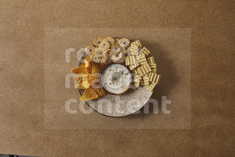 Assorted snacks on a pottery plate with a dipping on brown background