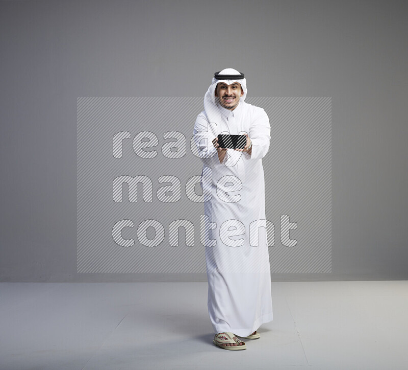 A Saudi man standing wearing thob and white shomag showing phone to camera on gray background