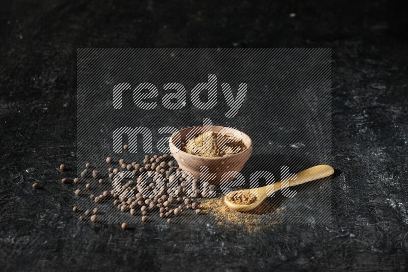 A wooden bowl and spoon full of allspice powder and whole balls spreaded on a black flooring