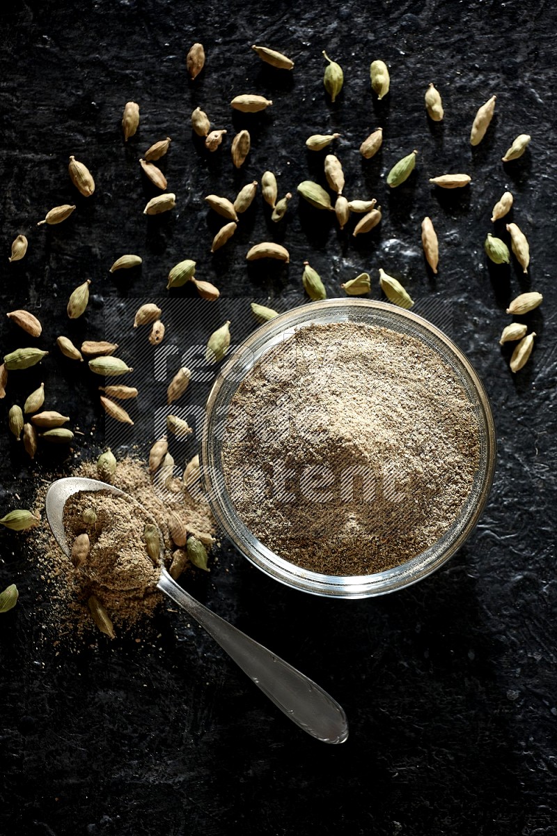 A glass bowl and a metal spoon full of cardamom powder with cardamom seeds beside them on textured black flooring