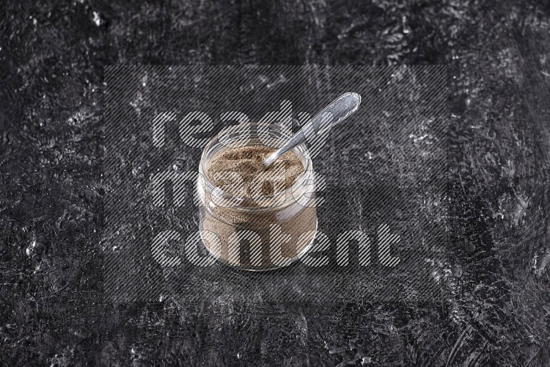 A glass jar full of black pepper powder and a metal spoon on a textured black flooring