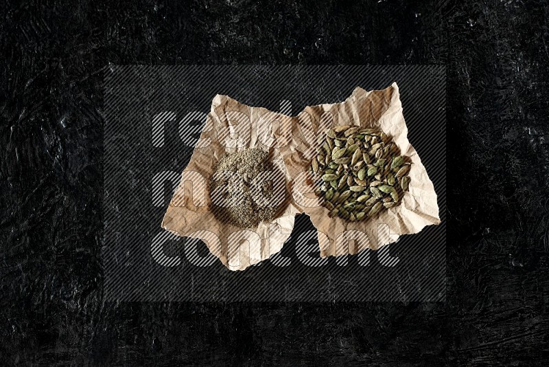 Cardamom seeds and cardamom powder in 2 crumpled pieces of paper on textured black flooring