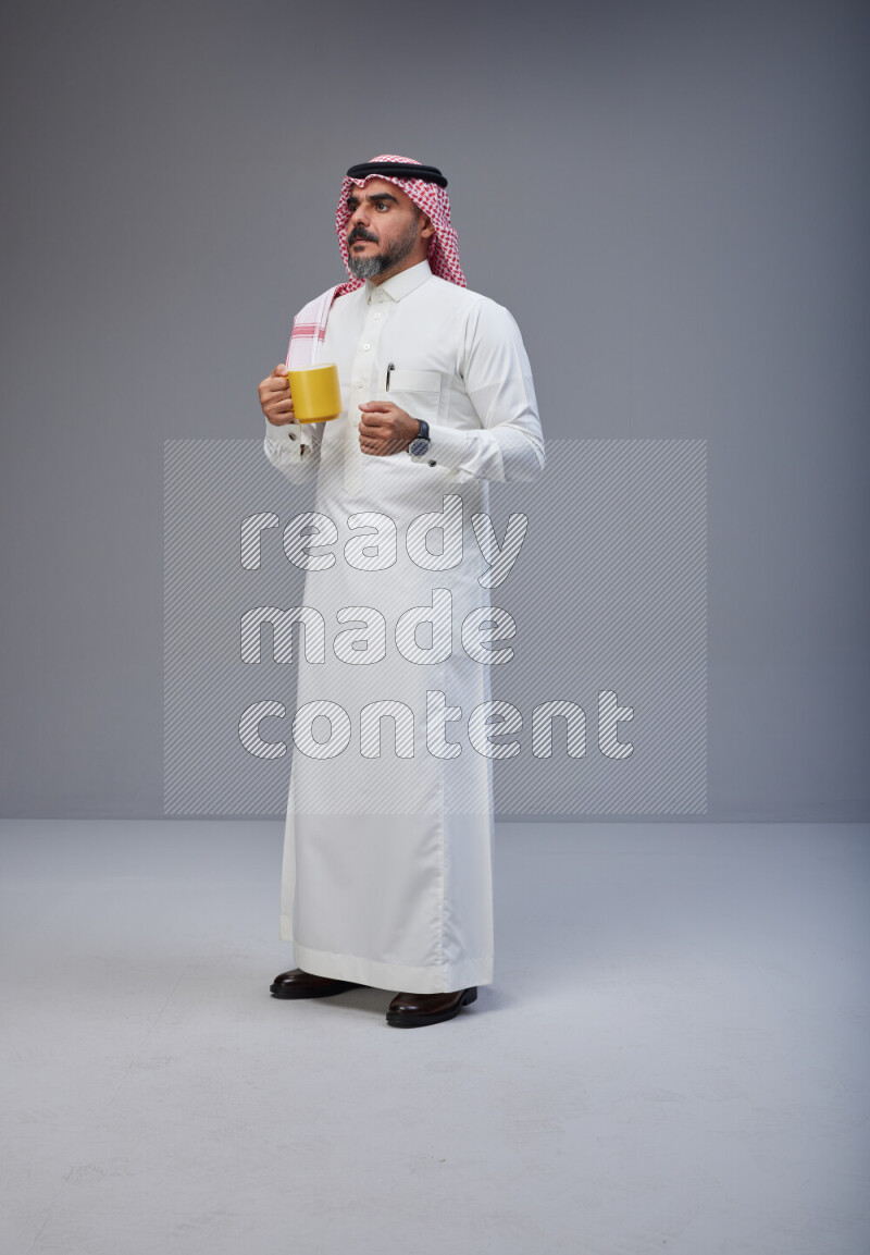 Saudi man Wearing Thob and red Shomag standing holding mug on Gray background