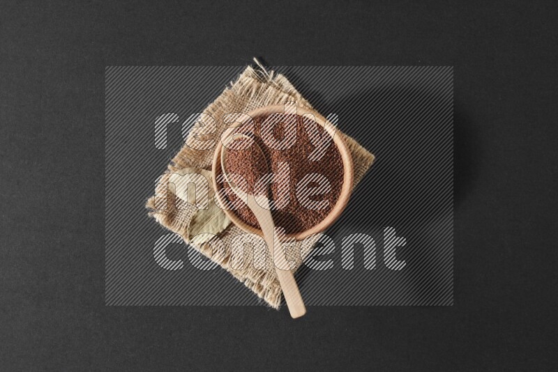 A wooden bowl full of garden cress seeds with wooden spoon full of the seeds on it on burlap fabric on a black flooring