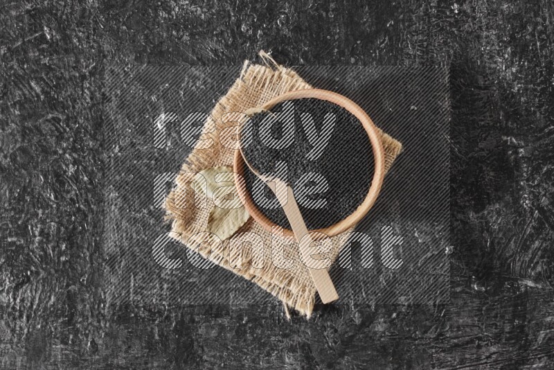 وعاء خشبي مع ملعقة خشبية ممتلئان ببذور حبة البركة علي قطعة من القماش علي خلفية سوداء