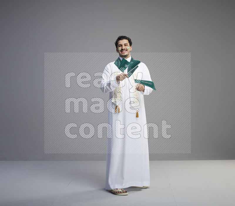 A Saudi man standing wearing thob and saudi flag scarf with face painting holding small Saudi flag on gray background