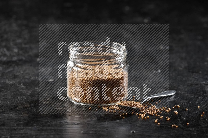 A glass jar and a metal spoon full of mustard seeds on a textured black flooring