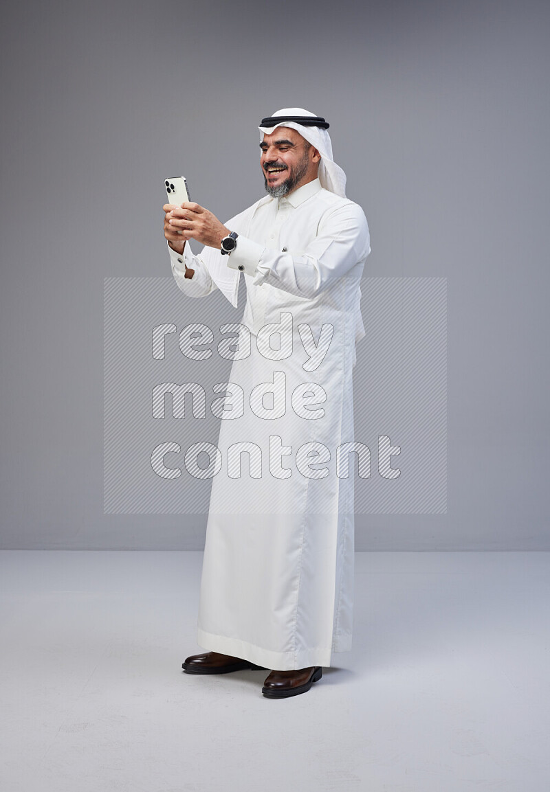 Saudi man Wearing Thob and white Shomag standing texting on phone on Gray background