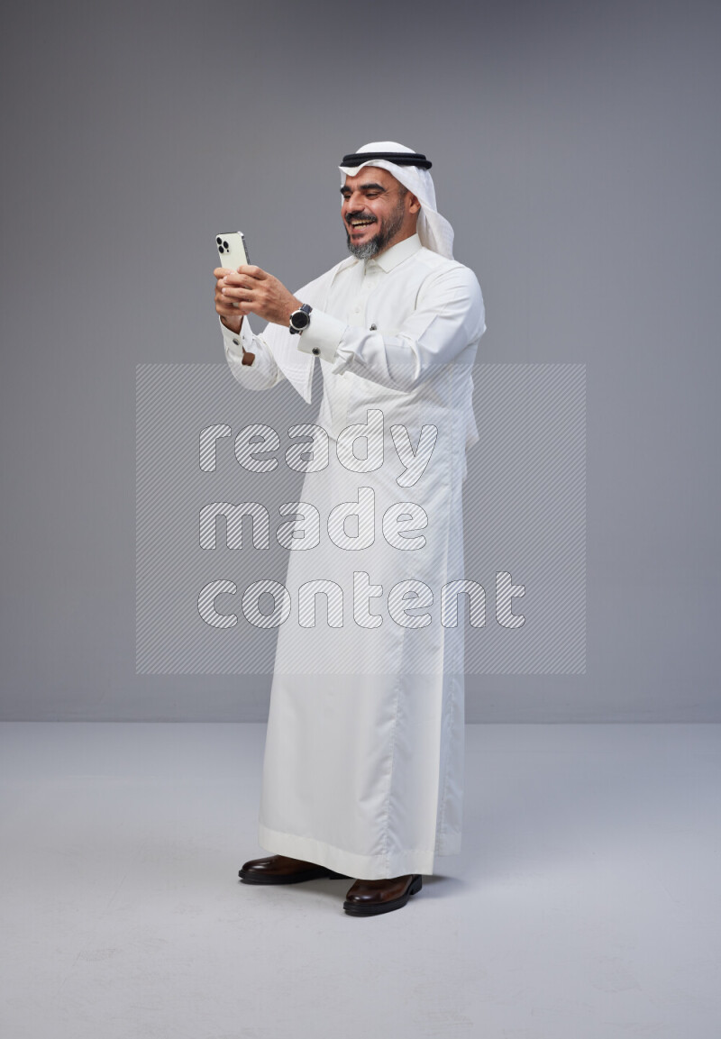 Saudi man Wearing Thob and white Shomag standing texting on phone on Gray background
