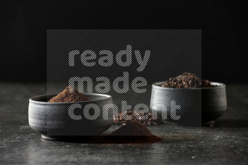 2 Black pottery bowls full of cloves and the other full of cloves powder on textured black flooring