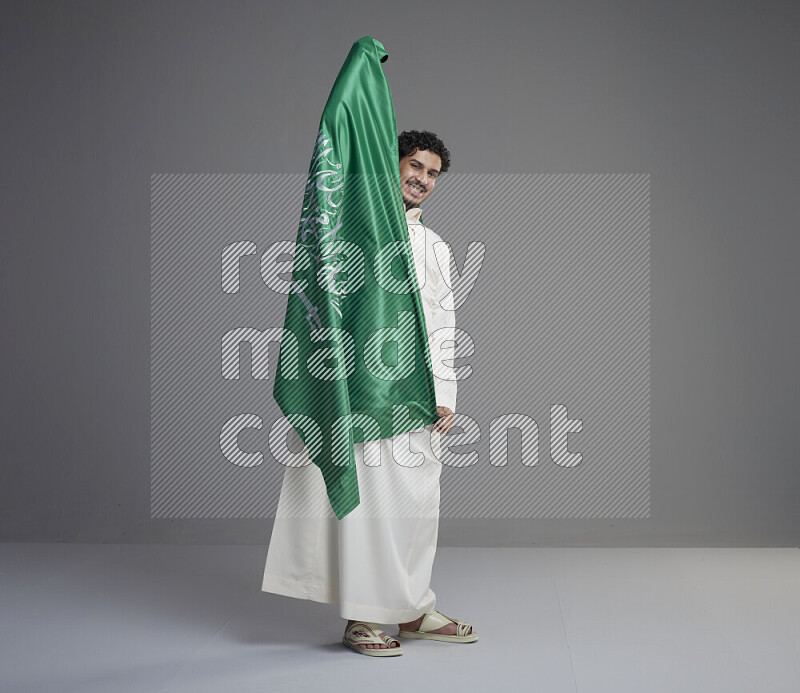 A saudi man standing wearing thob holding big saudi flag on gray background