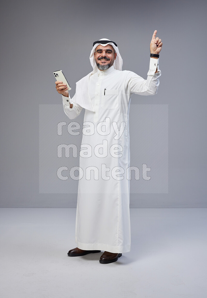 Saudi man Wearing Thob and white Shomag standing texting on phone on Gray background