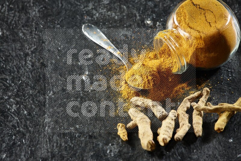A flipped glass spice jar and metal spoon full of turmeric powder and powder spilled out of it with dried whole fingers on textured black flooring