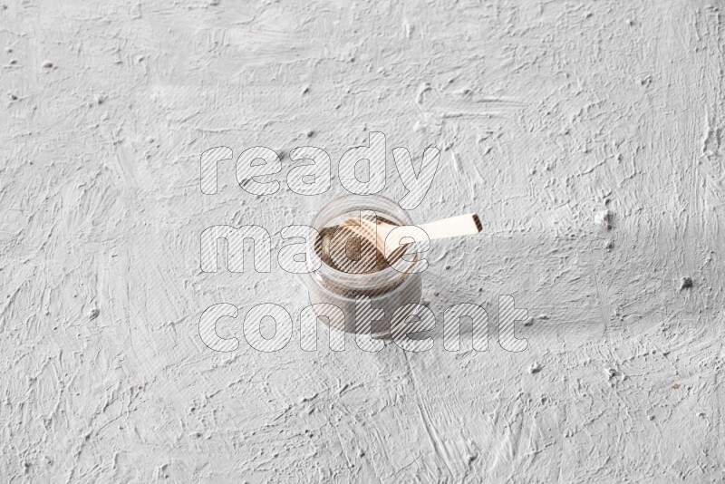 A glass jar full of black pepper powder with a wooden spoon on a textured white flooring