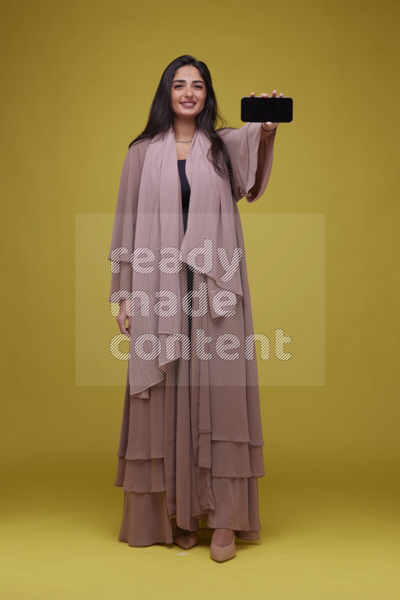 A woman Showing SmartPhone Screen on a Yellow Background wearing Brown Abaya