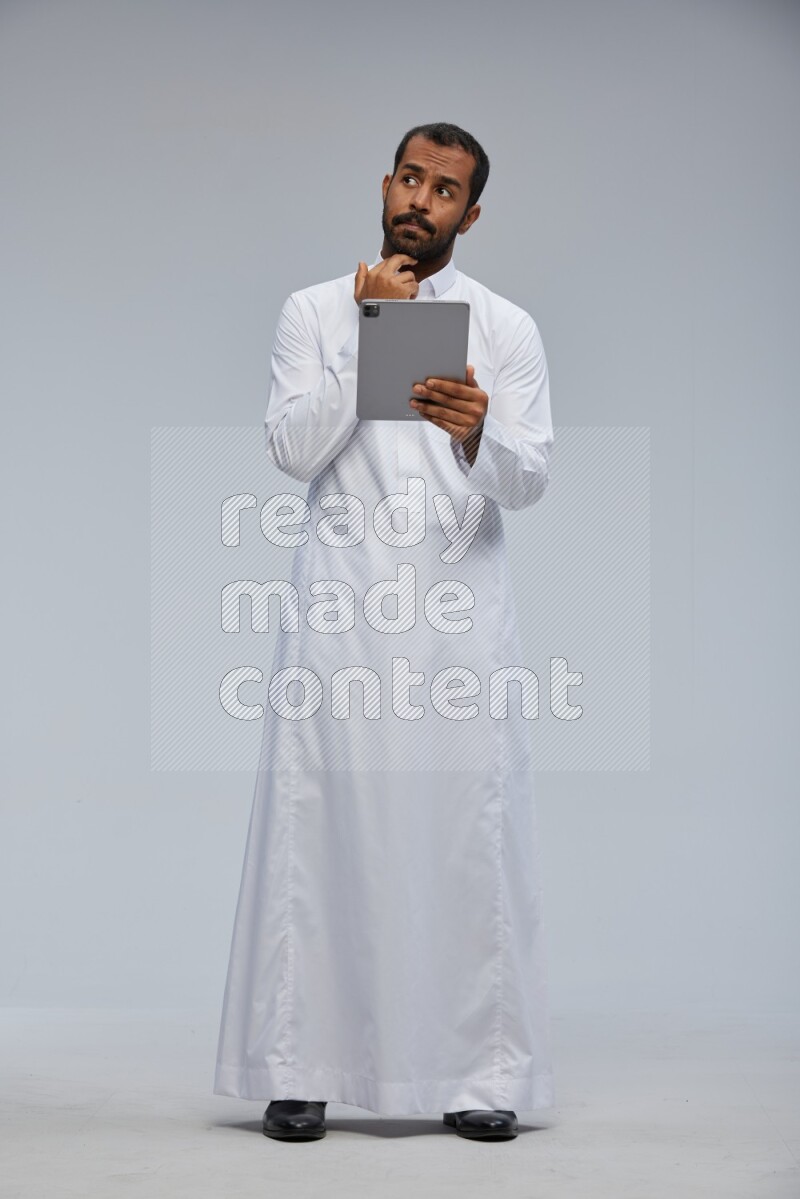 Saudi man wearing Thob standing working on tablet on Gray background