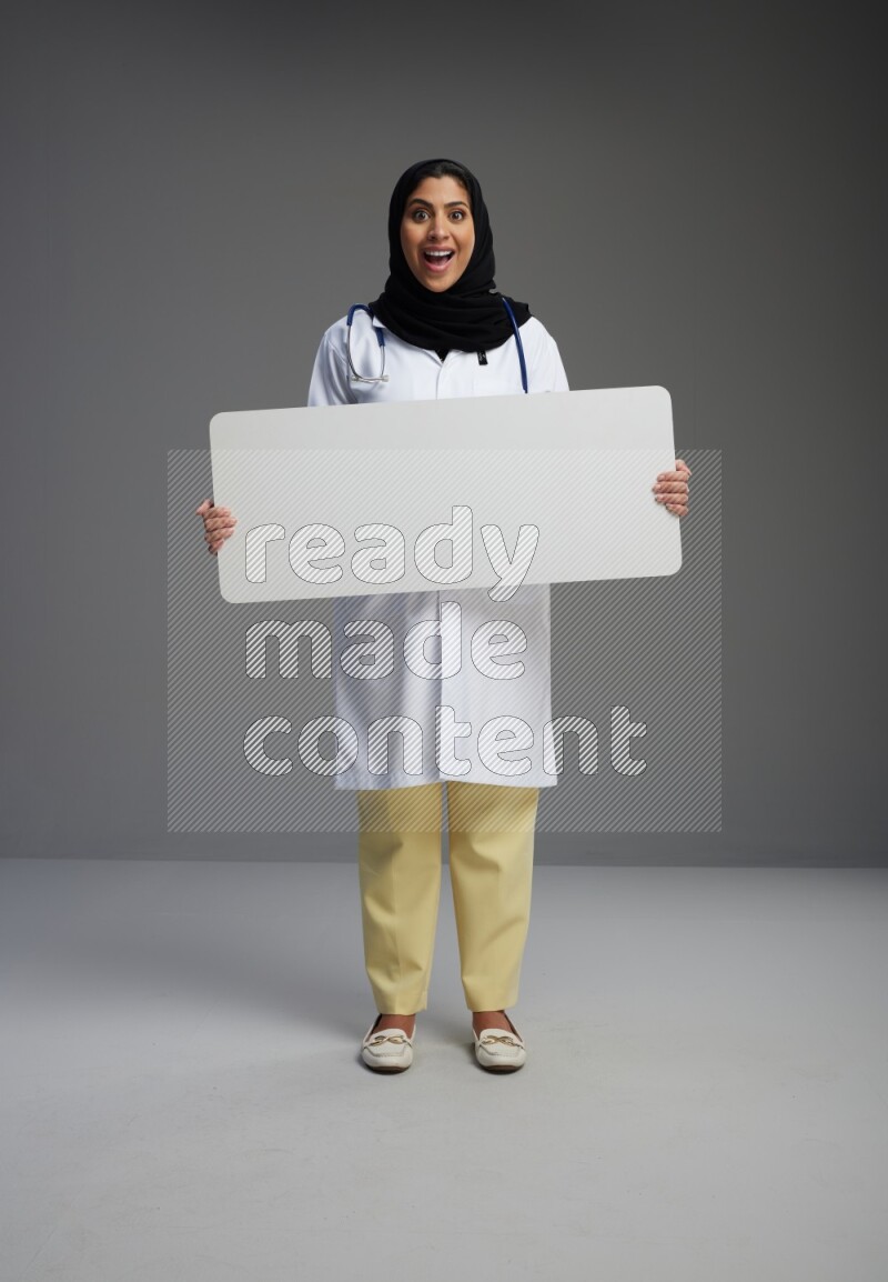 Saudi woman wearing lab coat with stethoscope standing holding board on Gray background